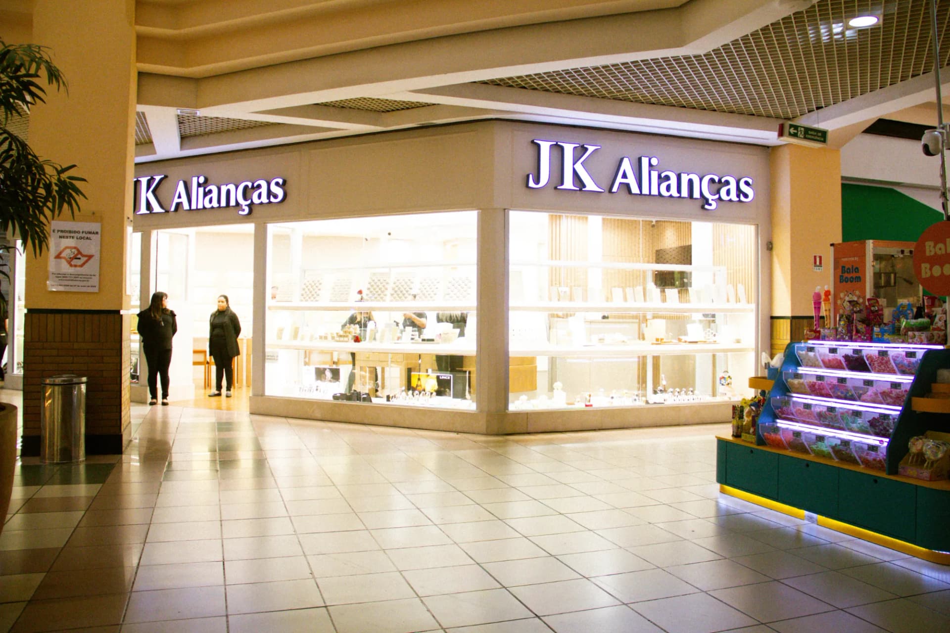 Vitrine iluminada da loja JK Alianças com letreiro branco, mostruário interno visível e duas atendentes na entrada